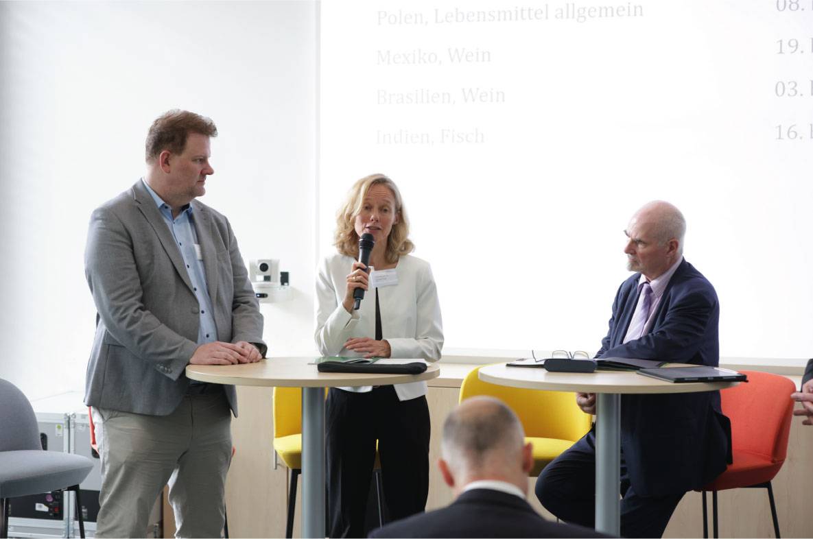 Diskussionsrunde auf dem 6. Außenwirtschaftsseminar der Agrar- und Ernährungswirtschaft in Bonn. Eine Referentin spricht mit Mikrofon zu den Herausforderungen und Chancen internationaler Agrar- und Ernährungsexporte. Zwei weitere Experten verfolgen die Diskussion an Stehtischen, während Fachbesucher im Publikum zuhören. Im Hintergrund sind Informationen zu internationalen Exportmärkten und Branchenthemen eingeblendet.