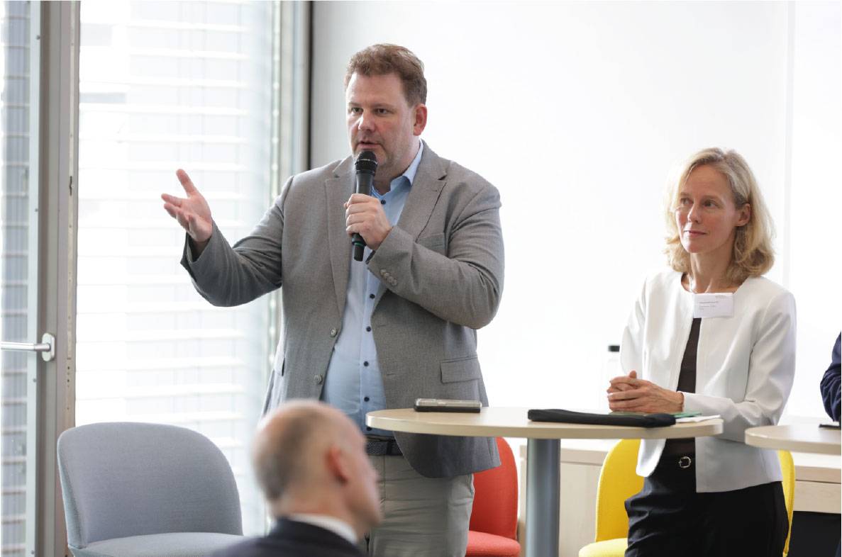 Experte spricht auf dem 6. Außenwirtschaftsseminar der Agrar- und Ernährungswirtschaft in Bonn zu aktuellen Entwicklungen im internationalen Agrar- und Ernährungshandel. Während seines Vortrags richtet er sich mit Mikrofon an die Teilnehmer der Veranstaltung. Eine weitere Referentin verfolgt die Diskussion am Stehtisch. Im Mittelpunkt stehen Exportmärkte, Handelsbeziehungen und die Wettbewerbsfähigkeit der deutschen Agrar- und Ernährungswirtschaft.