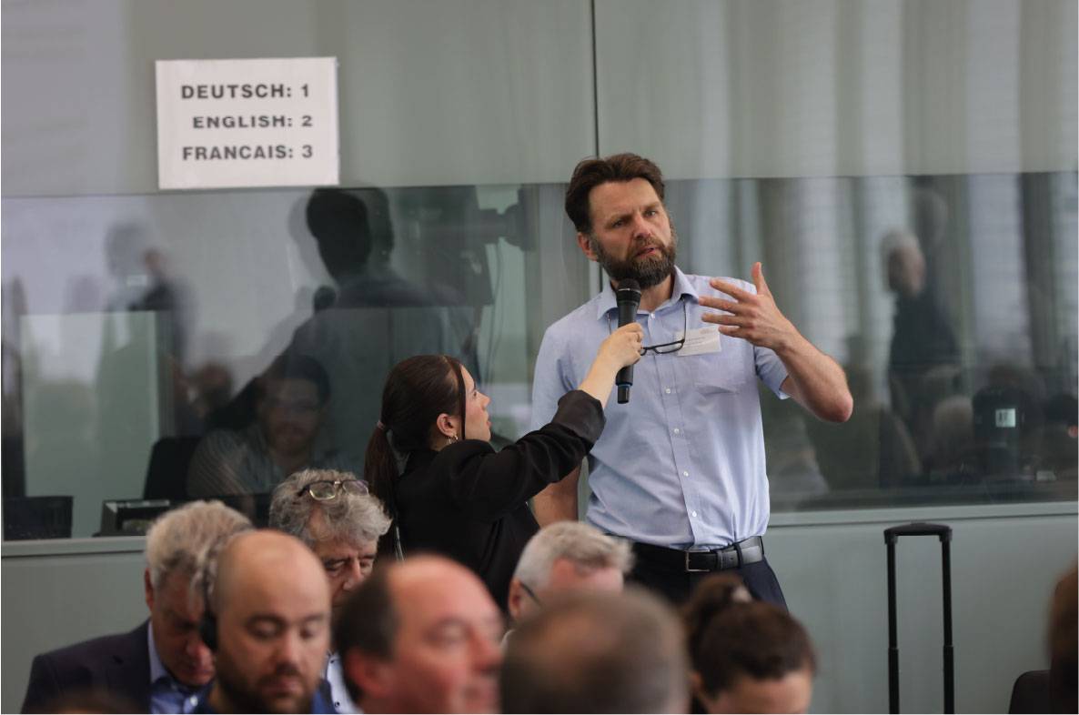 Ein Teilnehmer stellt während des 6. Außenwirtschaftsseminars der Agrar- und Ernährungswirtschaft in Bonn eine Frage aus dem Publikum. Eine Moderatorin hält ihm das Mikrofon, während zahlreiche internationale Gäste der Veranstaltung zuhören. Mehrsprachige Simultanübersetzung in Deutsch, Englisch und Französisch unterstützt den fachlichen Austausch zu aktuellen Themen des Agrar- und Ernährungsexports.