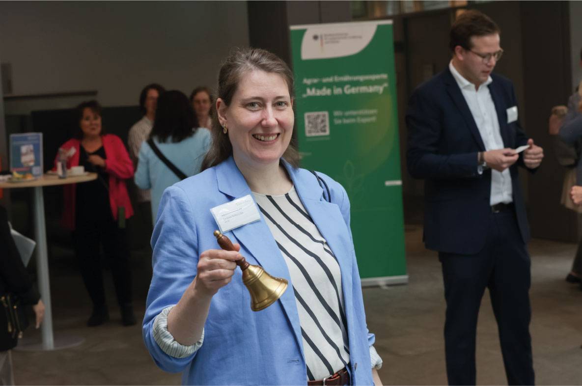 Teilnehmerin des 6. Außenwirtschaftsseminars der Agrar- und Ernährungswirtschaft in Bonn hält eine Handglocke während einer Networking-Pause in den Händen und lächelt in die Kamera. Im Hintergrund tauschen sich Gäste der Veranstaltung aus. Ein Banner der Exportförderinitiative „Made in Germany“ des Bundesministeriums für Landwirtschaft, Ernährung und Heimat verweist auf die Bedeutung internationaler Agrar- und Ernährungsexporte.