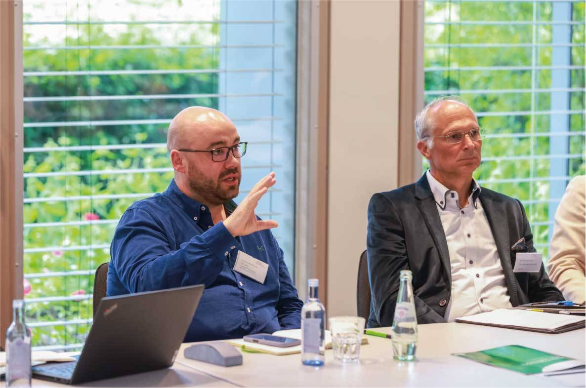 Teilnehmer des 6. Außenwirtschaftsseminars der Agrar- und Ernährungswirtschaft in Bonn diskutieren an einem Besprechungstisch über aktuelle Entwicklungen im internationalen Handel. Ein Teilnehmer erläutert seine Position mit einer Handbewegung, während weitere Fachvertreter aufmerksam zuhören. Die Veranstaltung fördert den fachlichen Austausch zu Exportmärkten, Außenwirtschaft und den Herausforderungen der deutschen Ernährungsindustrie.