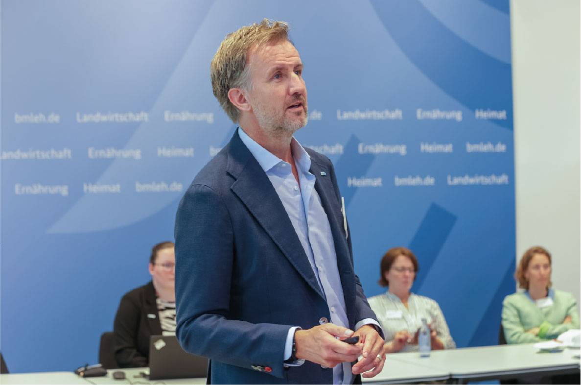 Referent hält einen Fachvortrag auf dem 6. Außenwirtschaftsseminar der Agrar- und Ernährungswirtschaft in Bonn. Vor einer Veranstaltungswand des Bundesministeriums für Landwirtschaft, Ernährung und Heimat präsentiert er aktuelle Entwicklungen im internationalen Agrar- und Lebensmittelhandel. Im Hintergrund verfolgen weitere Teilnehmer die Diskussion zu Exportmärkten, Außenwirtschaft und globalen Herausforderungen für die deutsche Ernährungsindustrie.