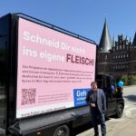 Christoph Minhoff, Hauptgeschäftsführer der Bundesvereinigung der Deutschen Ernährungsindustrie (BVE), steht vor einem schwarzen Werbefahrzeug mit einer großen pinkfarbenen Plakatwand der Europawahlkampagne „Geh wählen“. Darauf steht der Slogan „Schneid Dir nicht ins eigene FLEISCH!“. Im Hintergrund ist das Holstentor in Lübeck zu sehen.
