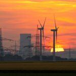 Sonnenuntergang hinter Windrädern, Hochspannungsleitungen und einem Kohlekraftwerk. Die Szene zeigt den Kontrast zwischen erneuerbarer und fossiler Energieerzeugung mit rauchenden Kühltürmen und modernen Windkraftanlagen im Vordergrund.