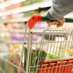 Nahaufnahme eines Einkaufswagens mit frischem Gemüse in einem Supermarkt. Eine Person mit grauem Pullover hält den grünen Griff des Wagens, während die Regale im Hintergrund leicht verschwommen sind.