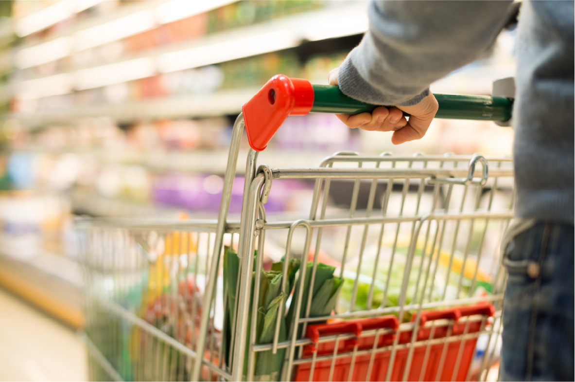 Nahaufnahme eines Einkaufswagens mit frischem Gemüse in einem Supermarkt. Eine Person mit grauem Pullover hält den grünen Griff des Wagens, während die Regale im Hintergrund leicht verschwommen sind.