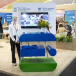 Messestand der Bundesvereinigung der Deutschen Ernährungsindustrie (BVE) auf der Anuga 2023. Im Vordergrund steht eine junge Frau am Empfangstresen in den Farben Blau und Grün mit dem BVE-Logo. Im Hintergrund sind weitere Informationsflächen, Sitzbereiche und bunte Infografiken zur Lebensmittelwirtschaft zu sehen.