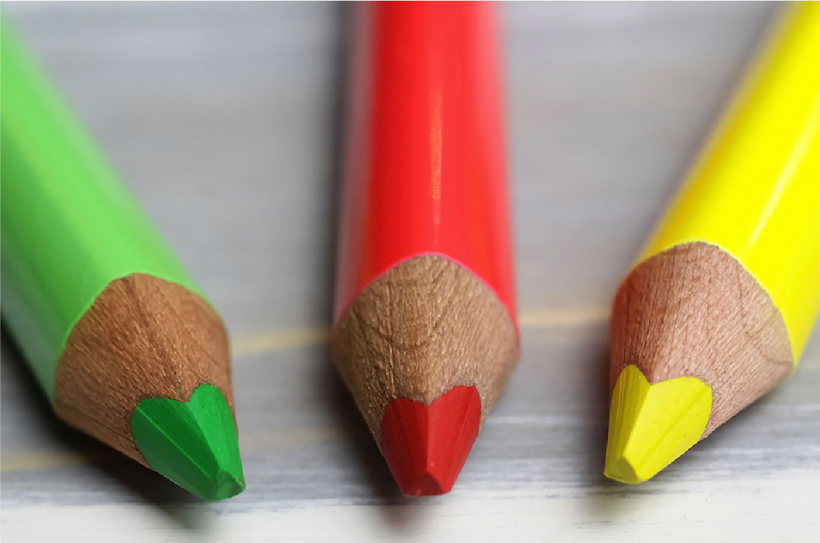 Drei dicke, angespitzte Buntstifte in den Farben Grün, Rot und Gelb liegen nebeneinander auf einem Tisch.