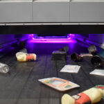 Sortieranlage mit violettem Licht, auf der verschiedene Kunststoffverpackungen auf einem Förderband liegen – Teil des Projekts „HolyGrail 2.0“. Ziel ist es, durch unsichtbare digitale Wasserzeichen auf Verpackungen die Recyclingquote zu erhöhen und die Qualität des Recyclings zu verbessern.
