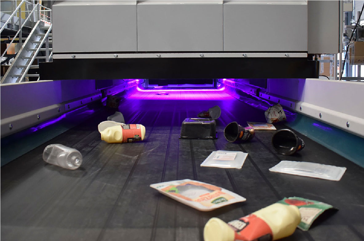 Sortieranlage mit violettem Licht, auf der verschiedene Kunststoffverpackungen auf einem Förderband liegen – Teil des Projekts „HolyGrail 2.0“. Ziel ist es, durch unsichtbare digitale Wasserzeichen auf Verpackungen die Recyclingquote zu erhöhen und die Qualität des Recyclings zu verbessern.