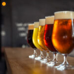 Fünf gefüllte Biergläser in einer Reihe auf einer Holztheke, jeweils mit unterschiedlich farbigem Bier – von dunkelbraun über bernsteinfarben bis hellgelb – und Schaumkrone. Im Hintergrund ist eine Bar mit dunklem, verschwommenem Interieur zu erkennen.