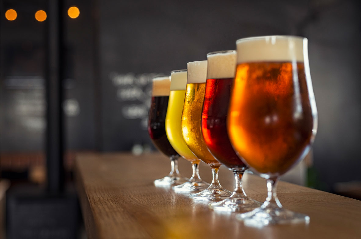 Fünf gefüllte Biergläser in einer Reihe auf einer Holztheke, jeweils mit unterschiedlich farbigem Bier – von dunkelbraun über bernsteinfarben bis hellgelb – und Schaumkrone. Im Hintergrund ist eine Bar mit dunklem, verschwommenem Interieur zu erkennen.
