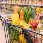 Einkaufswagen gefüllt mit frischem Obst, Gemüse und anderen Lebensmitteln in einem Supermarkt. Sichtbar sind unter anderem Bananen, Karotten, Gurken, Paprika, Lauch, Äpfel, eine Ananas, verschiedene Getränke und eine Flasche Öl. Im Hintergrund stehen gekühlte Regale mit weiteren Produkten.