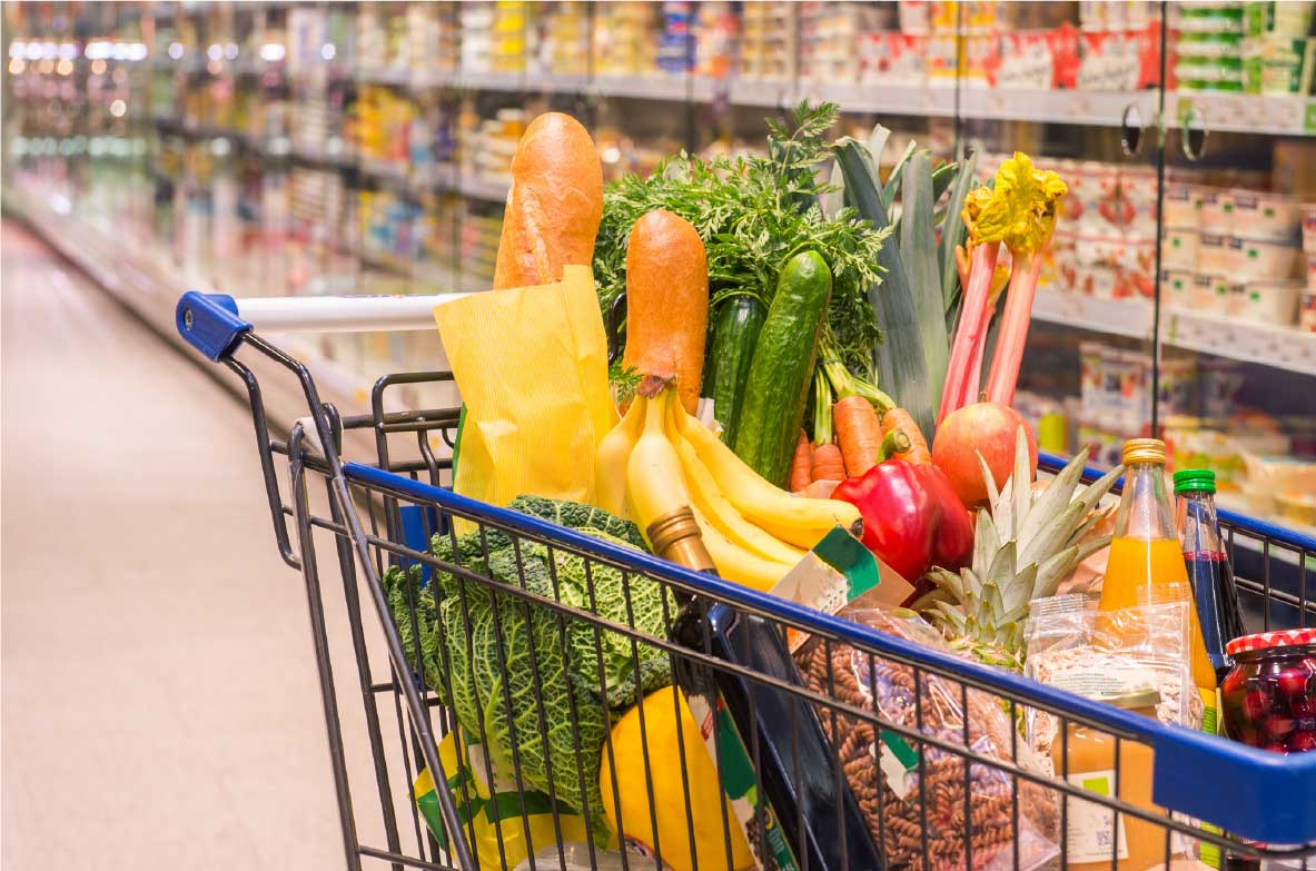 Einkaufswagen gefüllt mit frischem Obst, Gemüse und anderen Lebensmitteln in einem Supermarkt. Sichtbar sind unter anderem Bananen, Karotten, Gurken, Paprika, Lauch, Äpfel, eine Ananas, verschiedene Getränke und eine Flasche Öl. Im Hintergrund stehen gekühlte Regale mit weiteren Produkten.