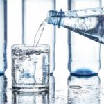 Nahaufnahme einer Wasserflasche, aus der kohlensäurehaltiges Wasser in ein Glas gegossen wird. Im Hintergrund stehen mehrere leere Plastikflaschen auf einer nassen Oberfläche.