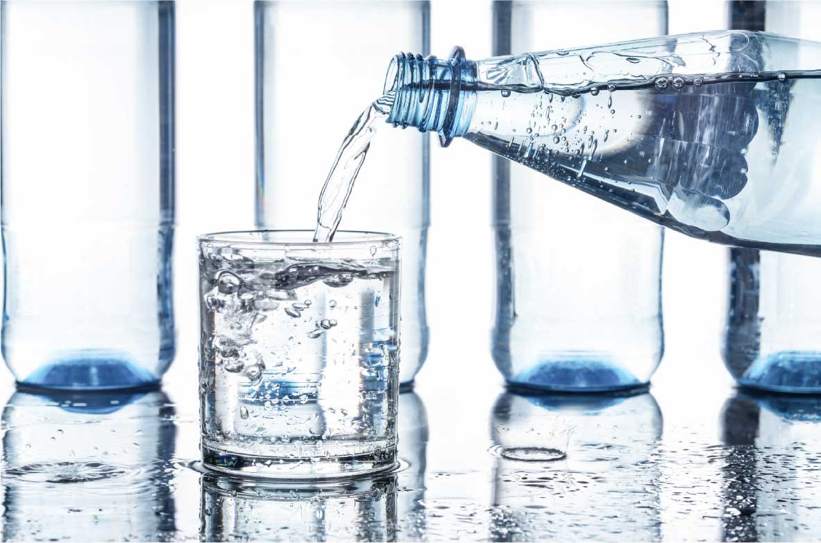 Nahaufnahme einer Wasserflasche, aus der kohlensäurehaltiges Wasser in ein Glas gegossen wird. Im Hintergrund stehen mehrere leere Plastikflaschen auf einer nassen Oberfläche.