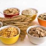 Verschiedene Schalen mit Frühstückscerealien, darunter Cornflakes, Haferflocken, Müsli und Weizenflakes, arrangiert neben Weizenähren auf einem weißen Hintergrund.