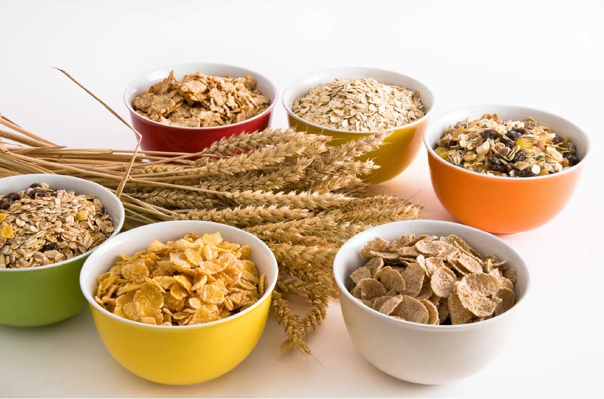 Verschiedene Schalen mit Frühstückscerealien, darunter Cornflakes, Haferflocken, Müsli und Weizenflakes, arrangiert neben Weizenähren auf einem weißen Hintergrund.