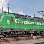 Grüne Elektro-Lokomotive der DB Cargo mit der Aufschrift 'Güter gehören auf die Schiene' auf einem Bahngleis. Die Oberleitung ist angeschlossen, im Hintergrund sind weitere Schienen und Strommasten zu sehen.