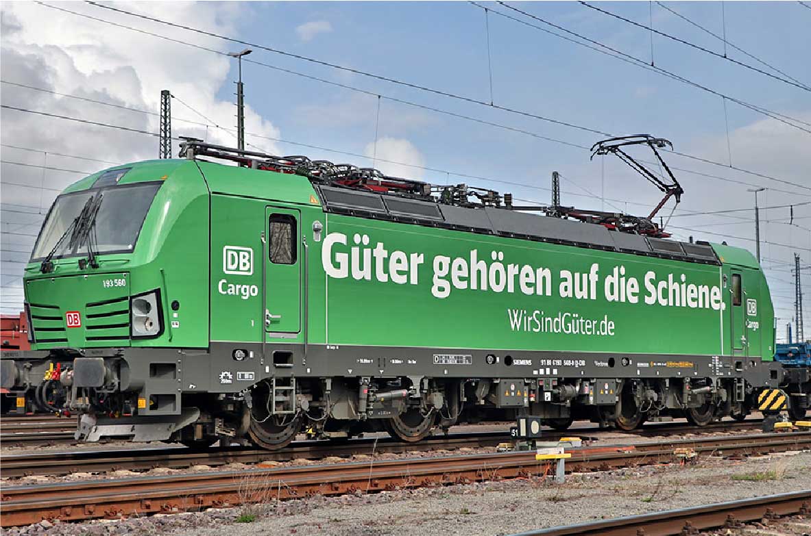 Grüne Elektro-Lokomotive der DB Cargo mit der Aufschrift 'Güter gehören auf die Schiene' auf einem Bahngleis. Die Oberleitung ist angeschlossen, im Hintergrund sind weitere Schienen und Strommasten zu sehen.