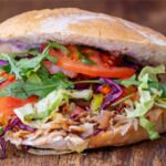 Nahaufnahme eines frisch zubereiteten Döners im aufgebackenen Fladenbrot, gefüllt mit geschnittenem Fleisch, Salat, Tomaten, Rotkohl, Rucola und einer Joghurtsoße. Der Döner liegt auf einer rustikalen Holzoberfläche.