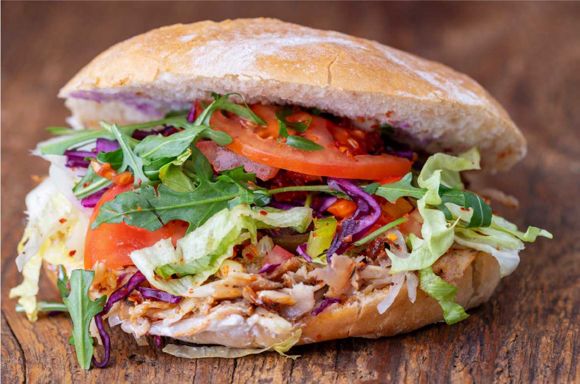 Nahaufnahme eines frisch zubereiteten Döners im aufgebackenen Fladenbrot, gefüllt mit geschnittenem Fleisch, Salat, Tomaten, Rotkohl, Rucola und einer Joghurtsoße. Der Döner liegt auf einer rustikalen Holzoberfläche.