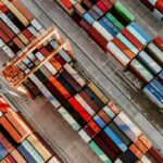 Luftaufnahme eines Containerhafens mit farbenfrohen, gestapelten Frachtcontainern und einem großen Verladekran. Die Container sind in langen Reihen auf dem Betonboden angeordnet, bereit zum Transport oder zur Weiterverladung.