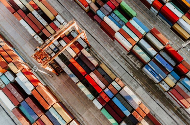 Luftaufnahme eines Containerhafens mit farbenfrohen, gestapelten Frachtcontainern und einem großen Verladekran. Die Container sind in langen Reihen auf dem Betonboden angeordnet, bereit zum Transport oder zur Weiterverladung.