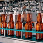 Braune Glasflaschen werden in einer Abfüllanlage automatisch befüllt. Die Flaschen stehen in Reih und Glied auf einem Förderband, während metallene Düsen von oben Flüssigkeit einfüllen.