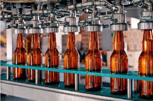 Braune Glasflaschen werden in einer Abfüllanlage automatisch befüllt. Die Flaschen stehen in Reih und Glied auf einem Förderband, während metallene Düsen von oben Flüssigkeit einfüllen.