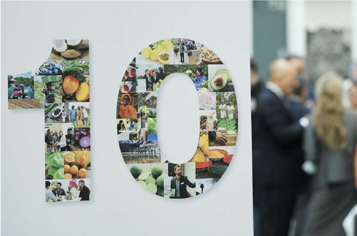 Große Zahl „10“, zusammengesetzt aus vielen kleinen Fotos von Lebensmitteln, Menschen in der Landwirtschaft und Handelsszenen. Die Zahl steht auf einer weißen Wand, im Hintergrund unterhalten sich mehrere Personen in Businesskleidung.