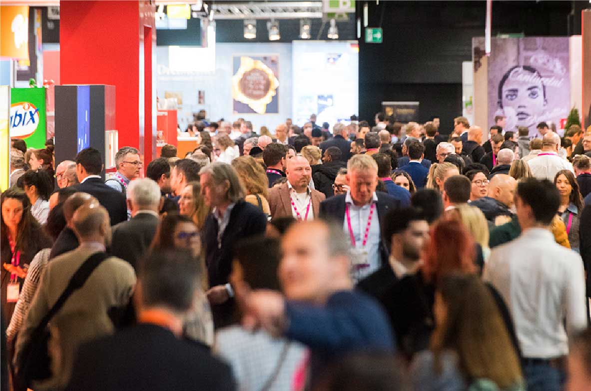 Menschenmenge auf der internationalen Süßwarenmesse ISM. Besucherinnen und Besucher bewegen sich durch die Messegänge, umgeben von farbenfrohen Ständen und Ausstellungsflächen. Die Szene zeigt das lebendige Treiben auf einer der größten Fachmessen für Süßwaren und Snacks.