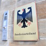 Eingang des Bundeskartellamts mit einem Schild an einer Steinwand. Das Schild zeigt einen schwarzen Bundesadler auf goldenem Hintergrund mit der Aufschrift 'Bundeskartellamt'. Daneben befindet sich eine Gegensprechanlage.