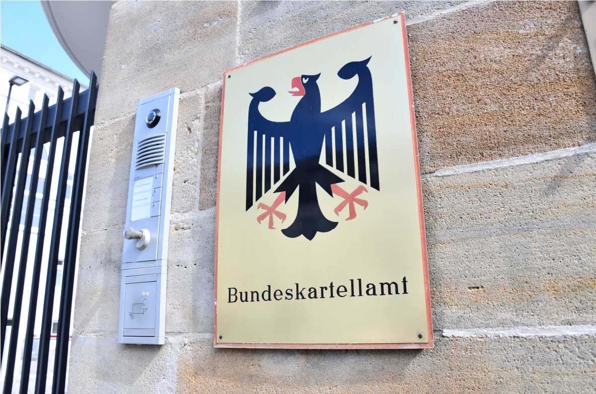 Eingang des Bundeskartellamts mit einem Schild an einer Steinwand. Das Schild zeigt einen schwarzen Bundesadler auf goldenem Hintergrund mit der Aufschrift 'Bundeskartellamt'. Daneben befindet sich eine Gegensprechanlage.