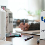 Mehrere individuell gestaltete Milchkartons stehen auf einem Tisch, verziert mit Namen, Zeichnungen und Logos. Im Hintergrund schreiben Personen in Notizbücher, während verschiedene Stifte auf dem Tisch liegen.