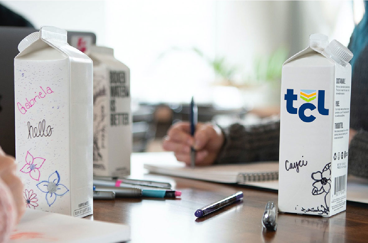 Mehrere individuell gestaltete Milchkartons stehen auf einem Tisch, verziert mit Namen, Zeichnungen und Logos. Im Hintergrund schreiben Personen in Notizbücher, während verschiedene Stifte auf dem Tisch liegen.