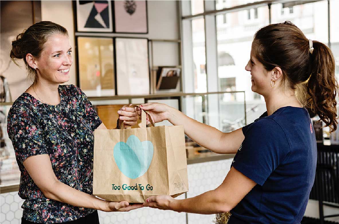 Zwei Frauen in einem Café tauschen eine braune Papiertüte mit dem Logo von 'Too Good To Go' aus. Eine Frau überreicht die Tüte, die andere nimmt sie mit einem Lächeln entgegen.