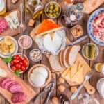 Ein reich gedeckter Holztisch mit einer Vielzahl unterschiedlicher Lebensmittel. Zu sehen sind verschiedene Käsesorten, Aufschnitt, Brot, Oliven, Tomaten, eingelegtes Gemüse, Nüsse, Dips sowie Bier- und Weingläser. Das Bild symbolisiert die Vielfalt an Lebensmitteln und kulinarischen Genüssen.