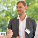 Max Webers, Co-Founder & CEO von COLIPI, steht in der Mitte des Bildes und unterhält sich mit zwei weiteren Personen. Er trägt ein graues Sakko, ein weißes Hemd und ein Namensschild. Im Hintergrund ist unscharf grüne Natur zu erkennen, das Gespräch findet offenbar im Außenbereich statt.