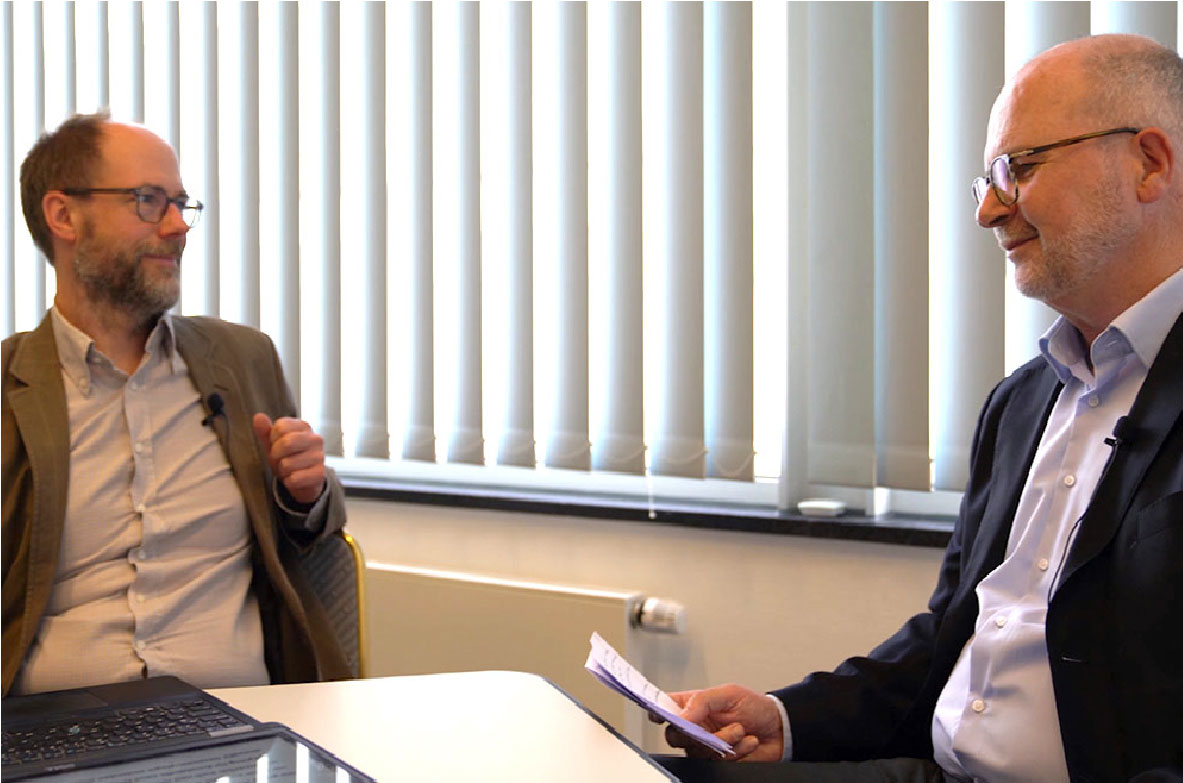 Zwei Männer im Gespräch bei einem Interview oder einer Diskussion. Rechts im Bild: Peter Feller, stellvertretender Hauptgeschäftsführer der BVE. Beide tragen Mikrofone und sitzen sich an einem Tisch mit Unterlagen gegenüber. Im Hintergrund vertikale Jalousien vor einem Fenster.