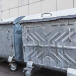 Reihe großer, metallener Müllcontainer mit Deckeln und Rollen, aufgestellt vor einer modernen Gebäudefassade. Die Behälter wirken industriell und sind für die Entsorgung großer Abfallmengen geeignet.
