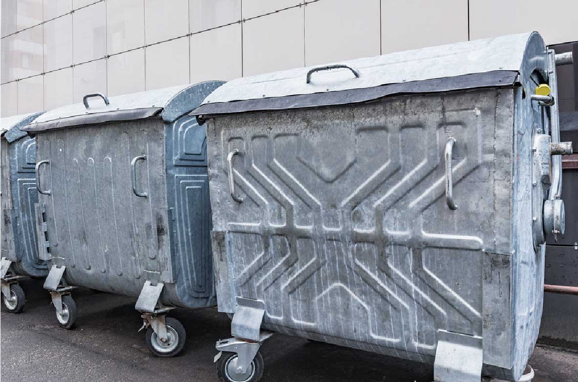 Reihe großer, metallener Müllcontainer mit Deckeln und Rollen, aufgestellt vor einer modernen Gebäudefassade. Die Behälter wirken industriell und sind für die Entsorgung großer Abfallmengen geeignet.