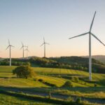 Mehrere Windräder stehen auf einem sanft hügeligen, grünen Feld. Im Hintergrund erstreckt sich eine bewaldete Landschaft bis zum Horizont unter einem klaren Himmel bei Sonnenuntergang.