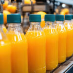 Eine Produktionsstraße auf der sich Ornagensaftflaschen aneinander reihen. Im Hintergrund frische Orangen und Saftpressen.