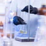 Eine Person im weißen Laborkittel und mit schwarzen Schutzhandschuhen führt in einem Labor eine präzise Flüssigkeitsmessung mit einer Pipette in ein Erlenmeyerkolbengefäß durch. Die Szene symbolisiert den biotechnologischen Prozess der Präzisionsfermentation. Im Hintergrund stehen unscharfe Laborflaschen und Geräte.
