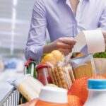 Person im Supermarkt hält einen Kassenbon in der Hand, während ein voller Einkaufswagen mit Lebensmitteln wie Gemüse, Getränken, Pasta und Brot vor ihr steht.