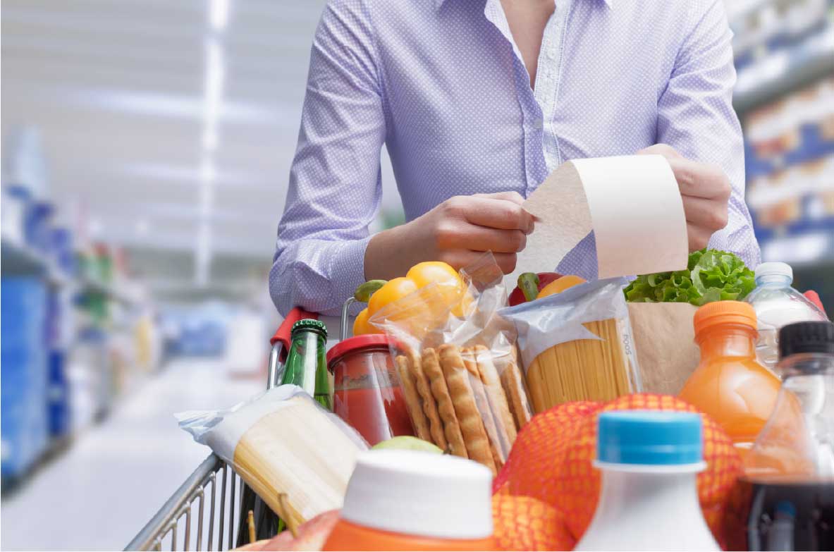 Person im Supermarkt hält einen Kassenbon in der Hand, während ein voller Einkaufswagen mit Lebensmitteln wie Gemüse, Getränken, Pasta und Brot vor ihr steht.