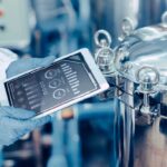 Eine Person in Schutzkleidung und blauen Handschuhen hält ein Tablet mit digitalen Anzeigen vor einen großen, silbernen Edelstahltank in einer industriellen Anlage. Auf dem Tablet sind grafische Messwerte zu Wasserstand, Druck und Temperatur zu sehen.