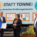 Drei Personen stehen vor einem bunt gestalteten Lkw mit der Aufschrift 'Tafel statt Tonne! Eine Aktion der deutschen Ernährungsindustrie'. Der Mann links hält eine geöffnete blaue Mappe mit Dokumenten, während die anderen beiden Personen mit ihm sprechen.