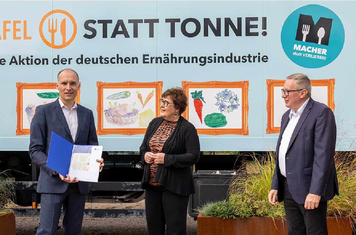 Drei Personen stehen vor einem bunt gestalteten Lkw mit der Aufschrift 'Tafel statt Tonne! Eine Aktion der deutschen Ernährungsindustrie'. Der Mann links hält eine geöffnete blaue Mappe mit Dokumenten, während die anderen beiden Personen mit ihm sprechen.